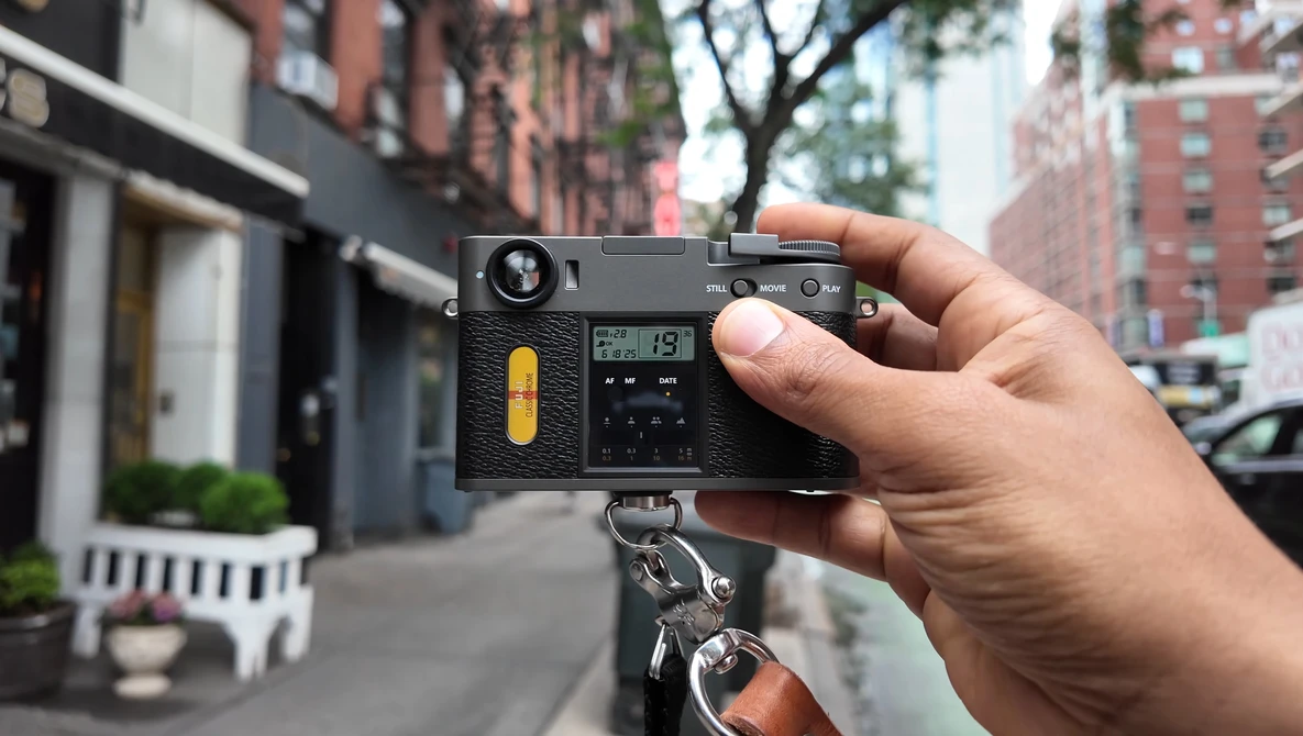 Hand holding a rangefinder film camera with yellow accent on the body, mounted on a tripod in an urban street setting.