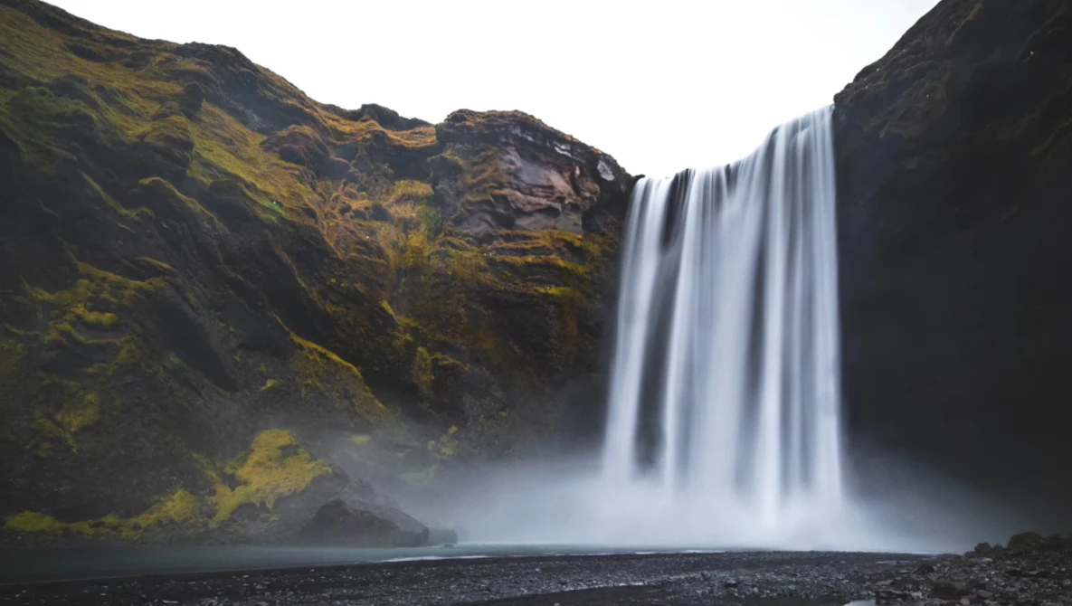 Waterfall cascading down dark basalt cliffs with moss-covered volcanic rock and mist rising from the pool below.
