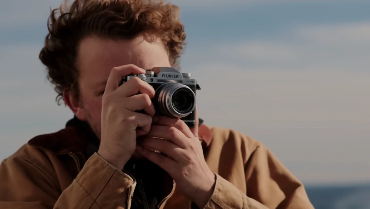 Person holding a rangefinder film camera up to their face, photographing against a coastal backdrop.