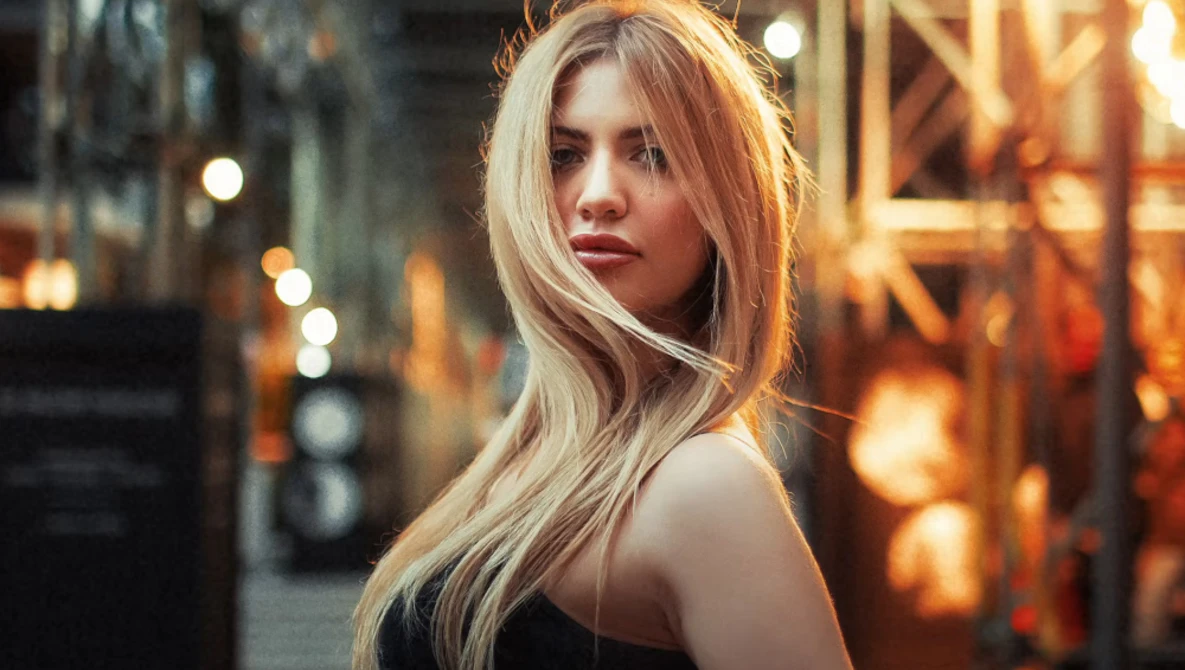 Woman with long blonde hair looking over shoulder in urban evening setting with warm golden lighting.