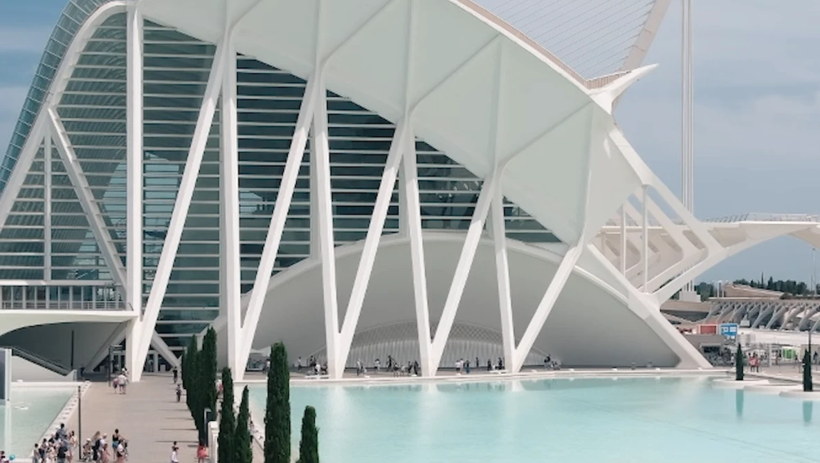 Modern white architectural structure with curved ribbed exterior beside turquoise reflecting pool, cypress trees, and glass building facade.