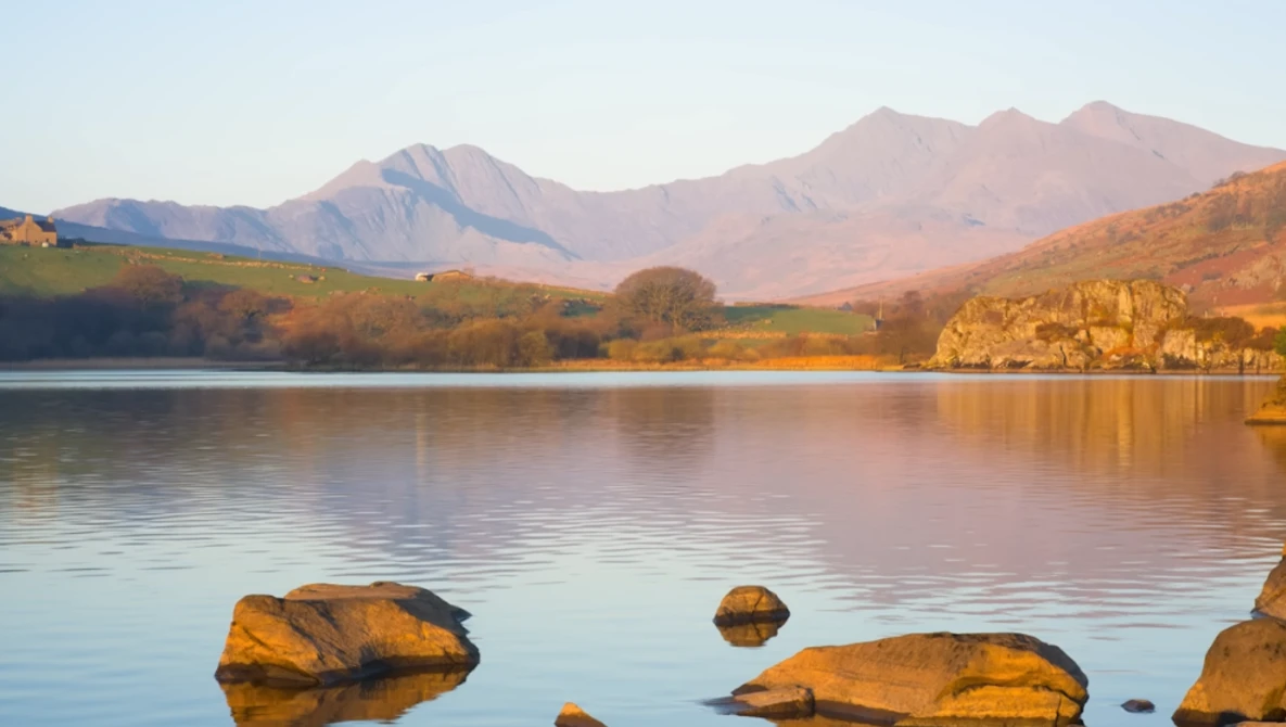 Mountain lake at golden hour with foreground rocks and tree-lined shoreline under clear sky.