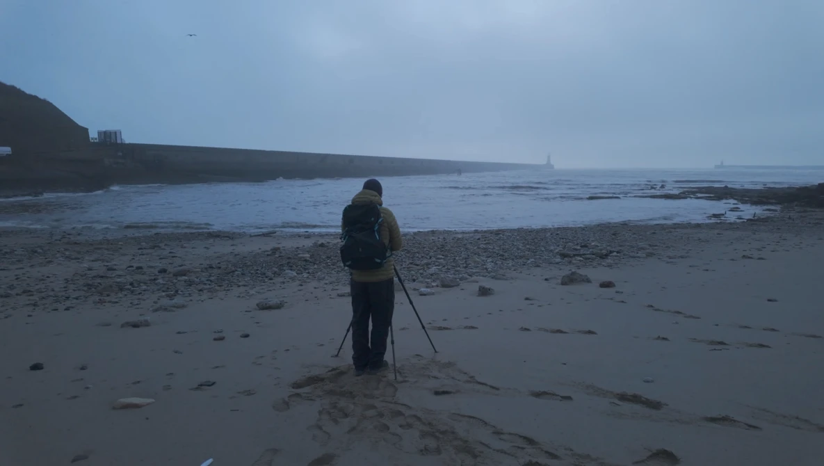 Photographer with tripod standing on wet beach at dusk, facing calm water and distant breakwater.