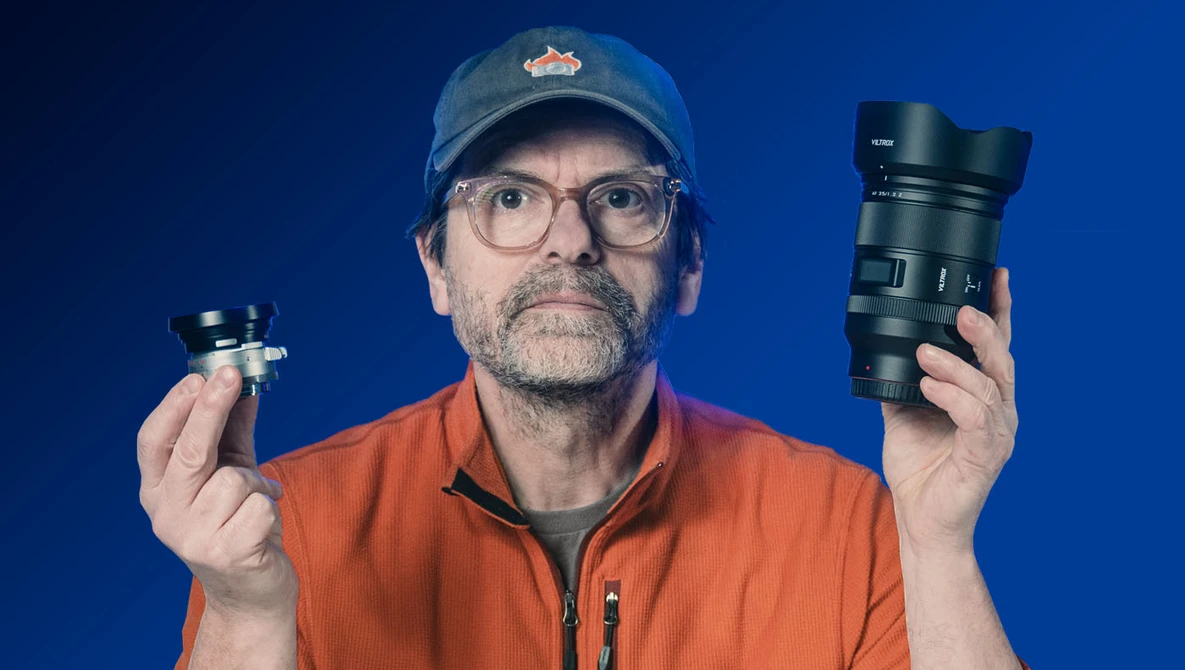 Photographer holding a compact prime lens in one hand and a larger telephoto lens in the other against a blue background.