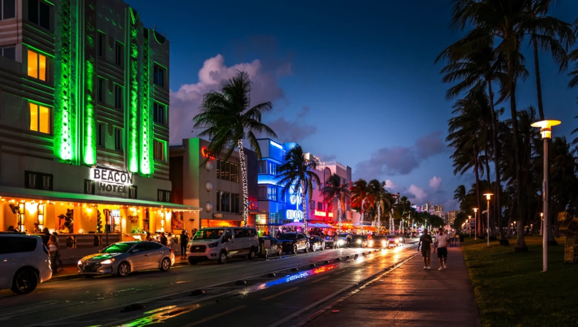 Art Deco hotel facades illuminated with neon green and blue lighting along a palm-lined street at dusk.