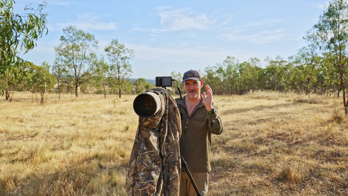 Wildlife photographer holding a telephoto lens in an open woodland savanna landscape.