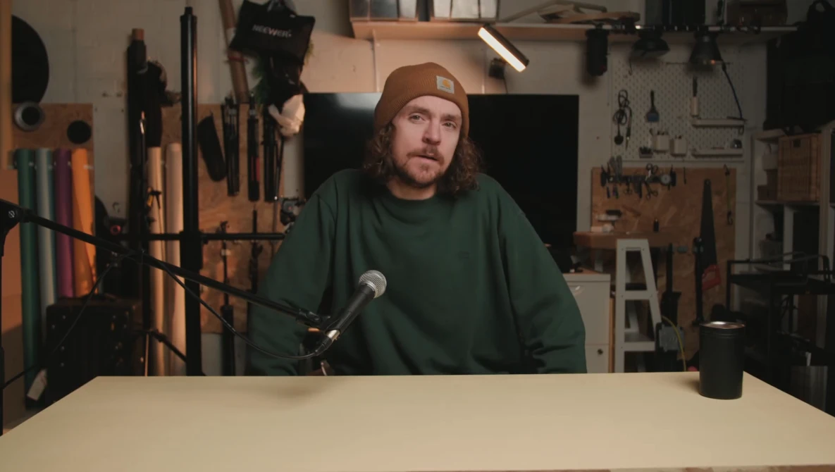 Man speaking into microphone at table in workshop with tools and lighting equipment in background.