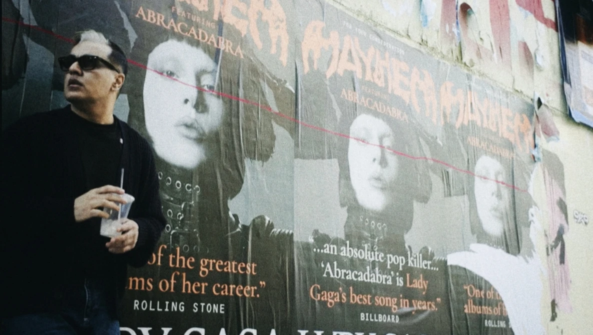 Man in sunglasses and black shirt standing in front of large promotional posters with black and white portrait imagery and text.