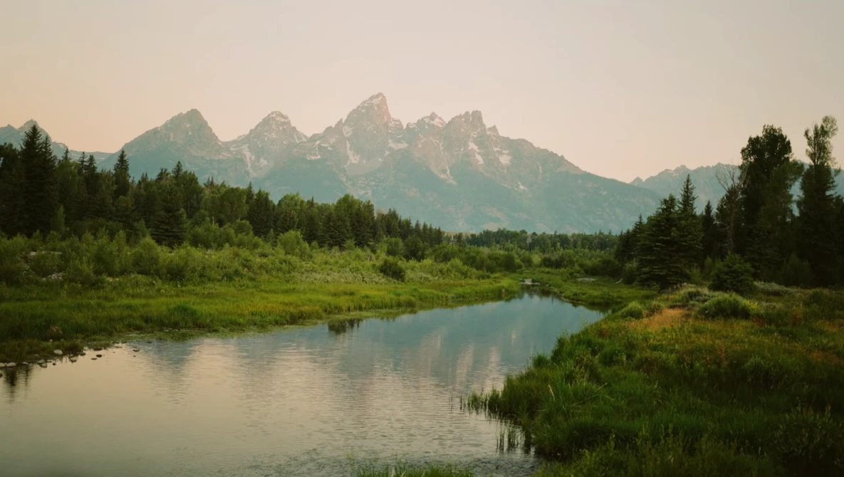 Mountain range with jagged peaks reflected in still river, surrounded by dense evergreen forest.