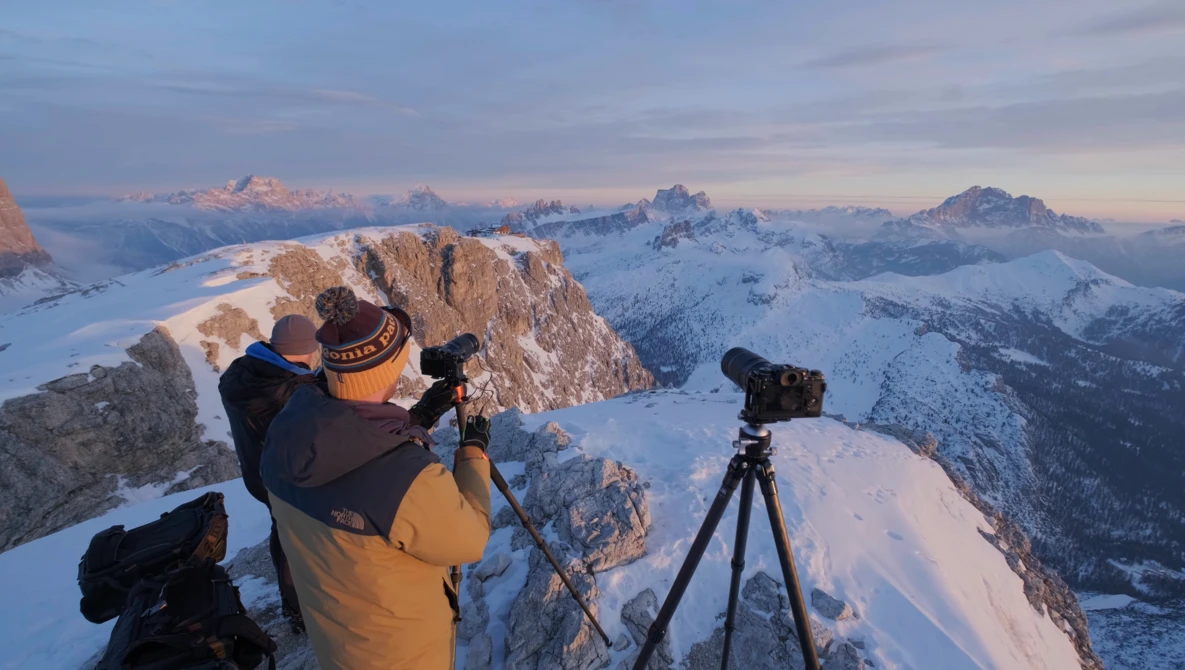 Photographer on snowy alpine ridge with camera on tripod overlooking snow-covered peaks at golden hour.