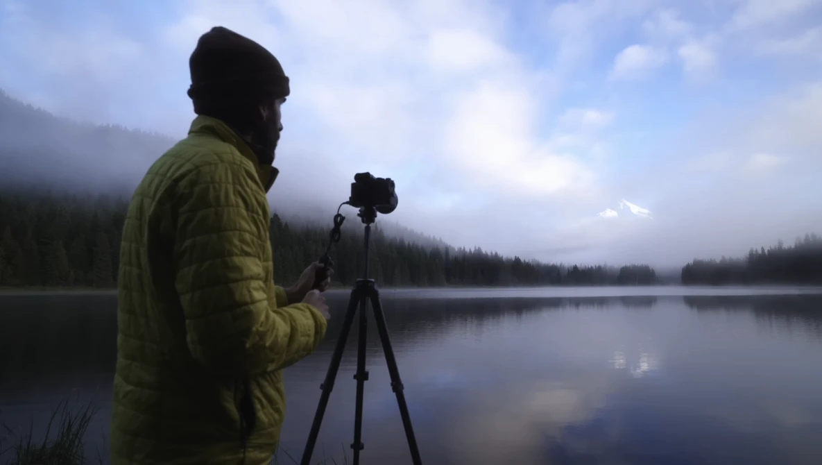 Photographer in yellow jacket operating tripod-mounted camera beside misty lake with forested shoreline.