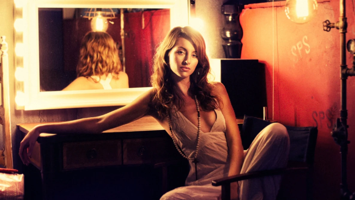 Woman in light-colored dress seated at illuminated vanity mirror in warm-toned studio setting.