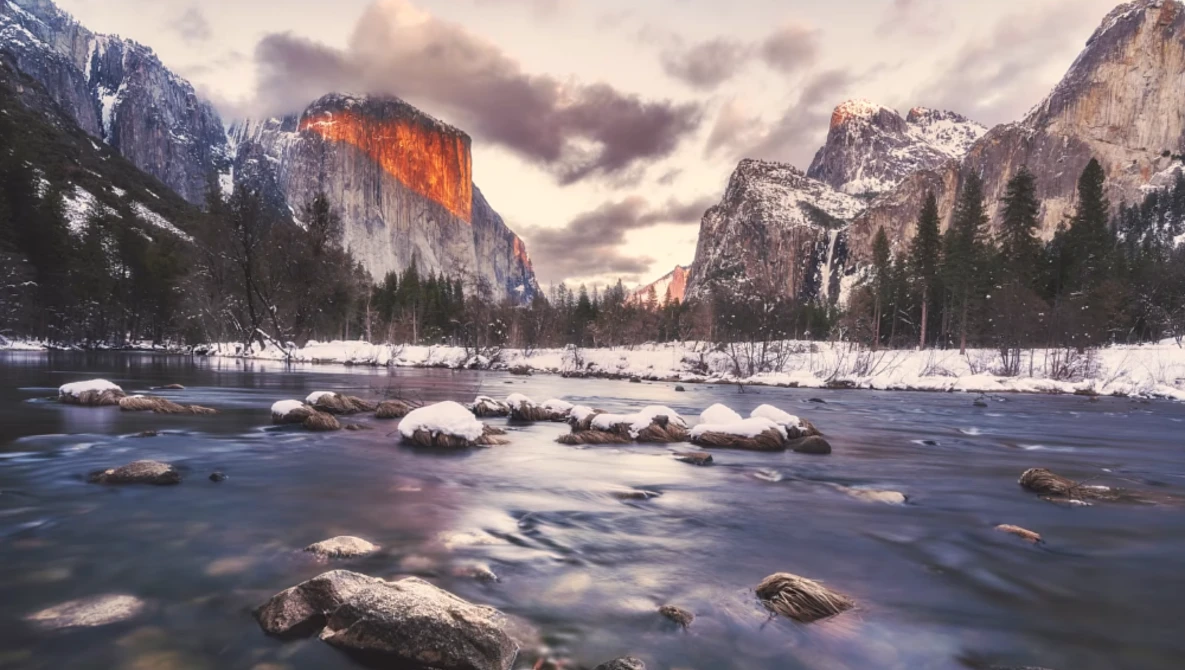 River flowing through a snowy valley with granite cliffs and evergreen forests under dramatic cloudy sky.