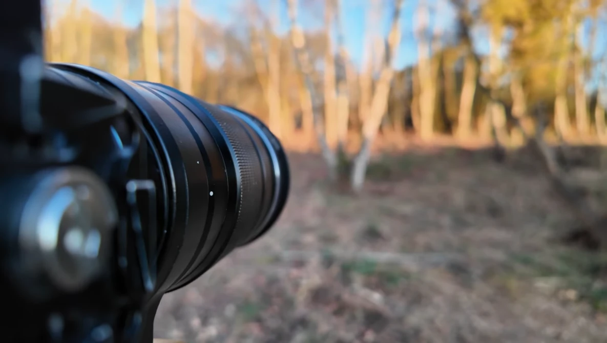 Camera lens in sharp focus with blurred architectural structures and autumn foliage in background.