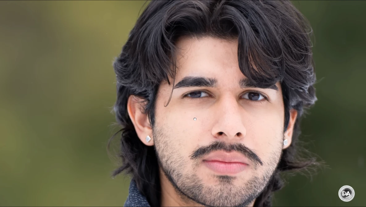 Portrait of a man with shoulder-length dark hair and facial hair against a soft green background.