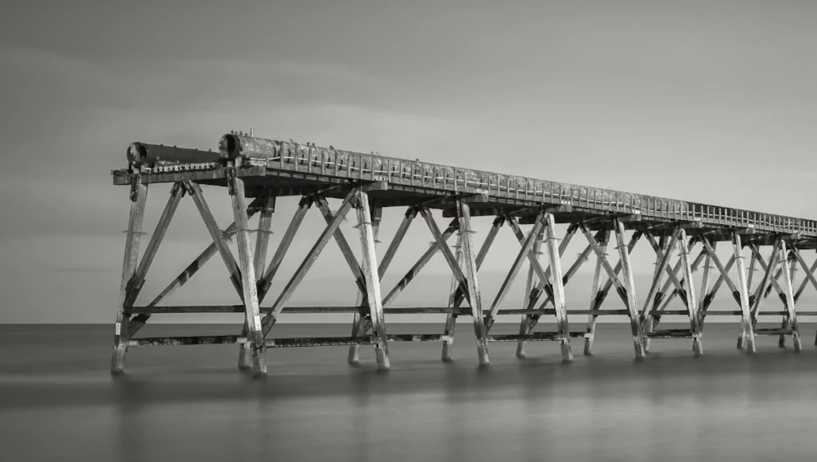 Long wooden pier with repetitive X-braced support structure extending into calm water under overcast sky.