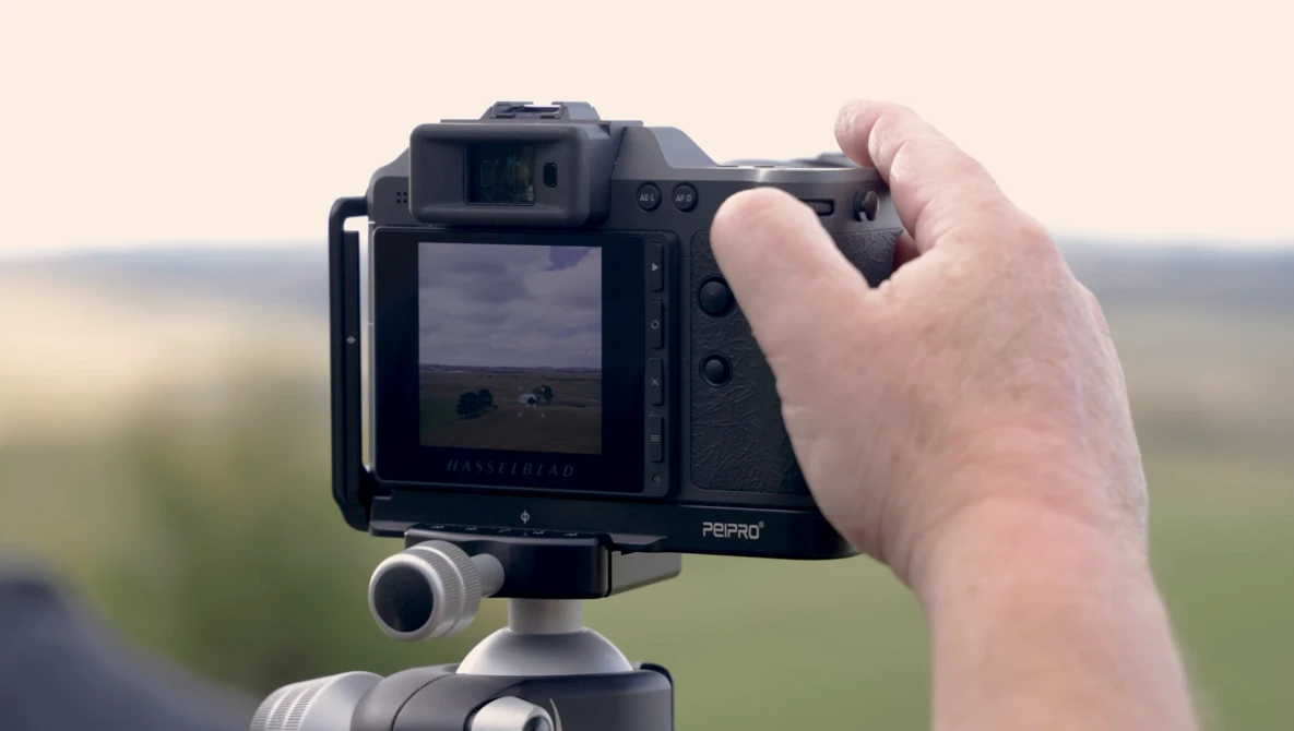 Photographer holding a medium format camera on a tripod, viewing the outdoor landscape through the LCD screen.
