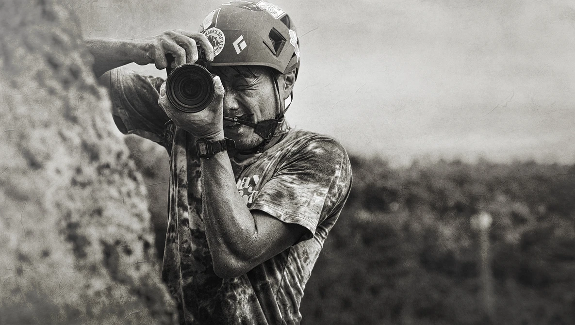 Climber in helmet peers through camera lens while secured to rock wall.