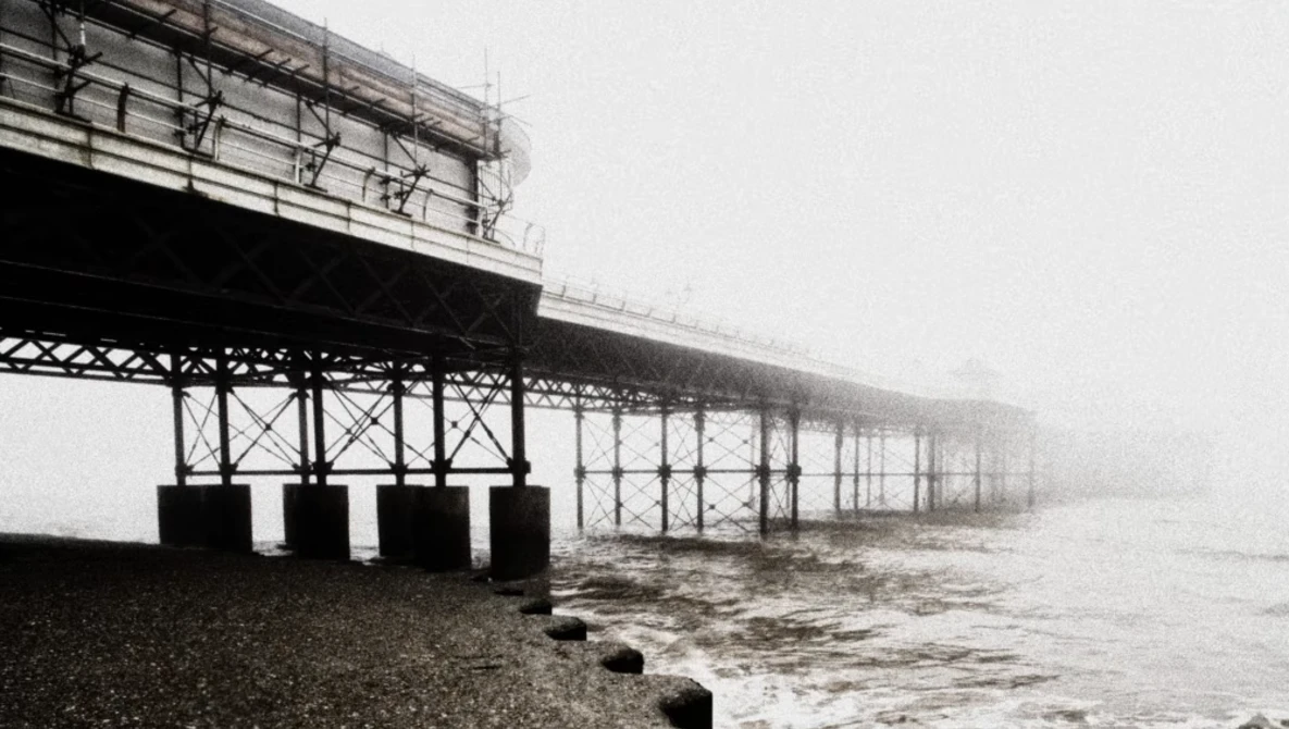 Pier structure receding into thick fog with dark metal framework and support pillars extending into turbulent water.
