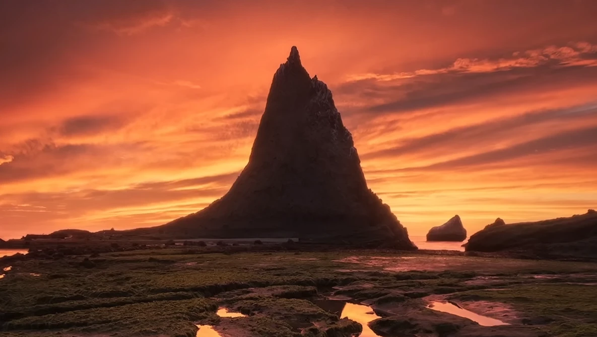 Dramatic silhouette of a tall sea stack against a vibrant orange and yellow sunset sky with reflection in wet tidal flats.