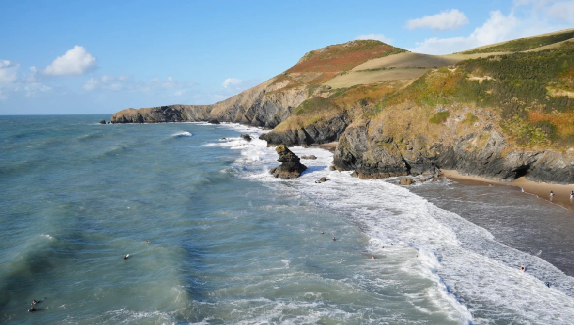 Rocky coastal cliffs with turquoise water and breaking waves beneath grassy headland.
