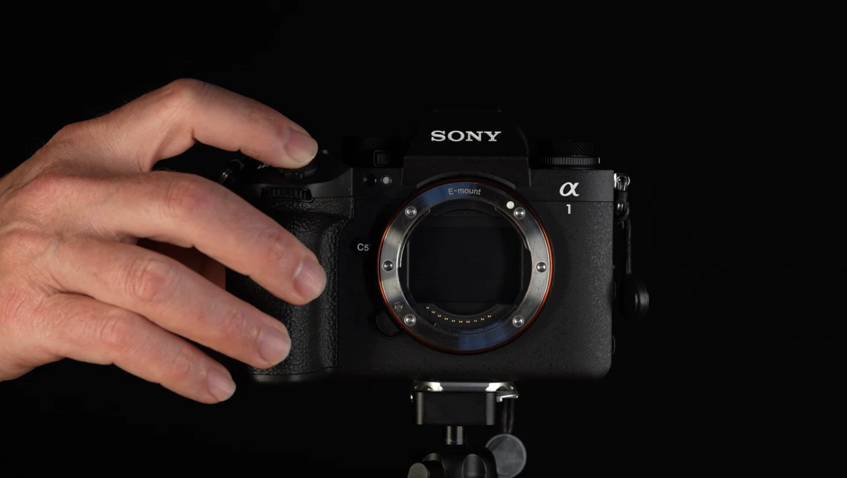 Hands holding a Sony Alpha mirrorless camera body without a lens attached, mounted on a tripod against a black background.
