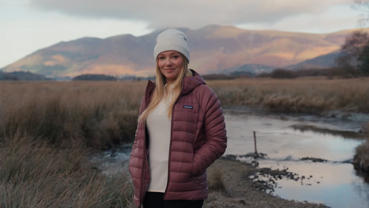 Woman in burgundy puffer jacket and white beanie standing in moorland landscape with stream and mountains behind.