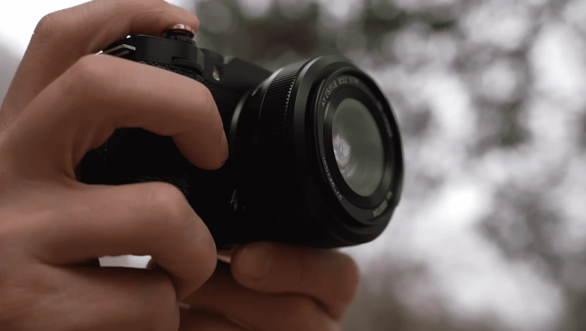Hands holding a compact mirrorless camera with a standard zoom lens, photographing outdoors.