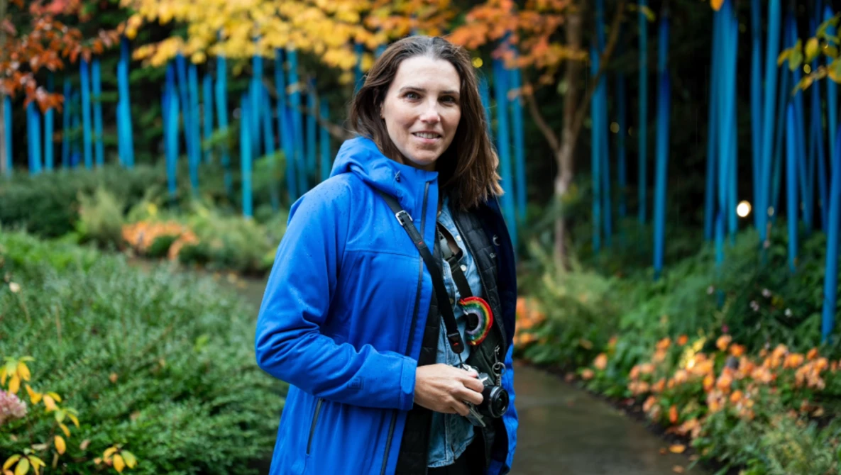 Woman holding a camera wearing a bright blue jacket in an autumn garden setting.