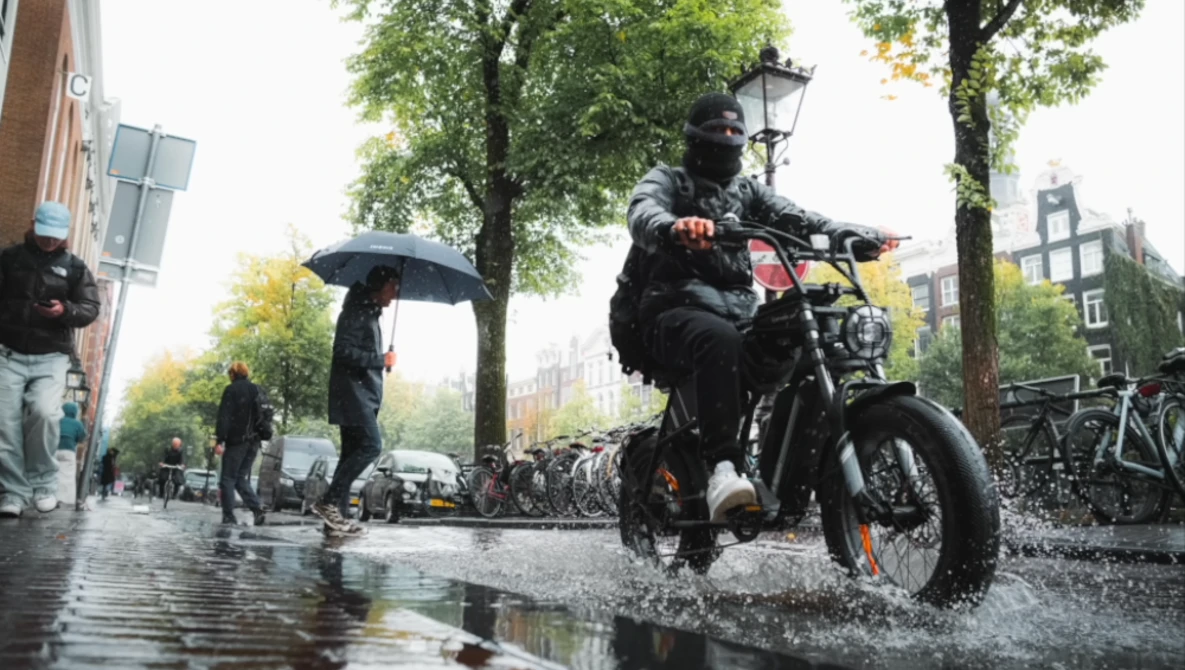 Man riding a motorcycle through a wet urban street during rain, splashing through water.