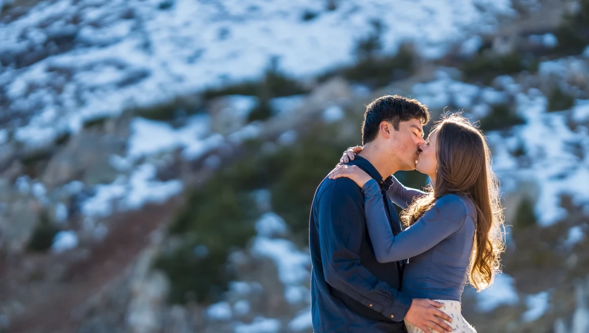 Couple kissing outdoors with snow-covered mountains and evergreen trees in soft golden hour light.