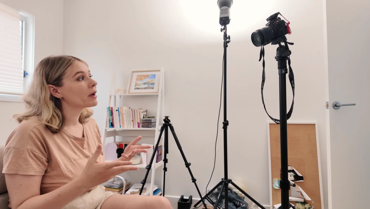 Woman sitting in a home studio setup, gesturing while speaking to camera during a recording session.