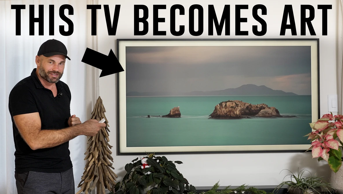 Man in black polo shirt pointing to framed landscape photo displaying turquoise water and rocky islands.