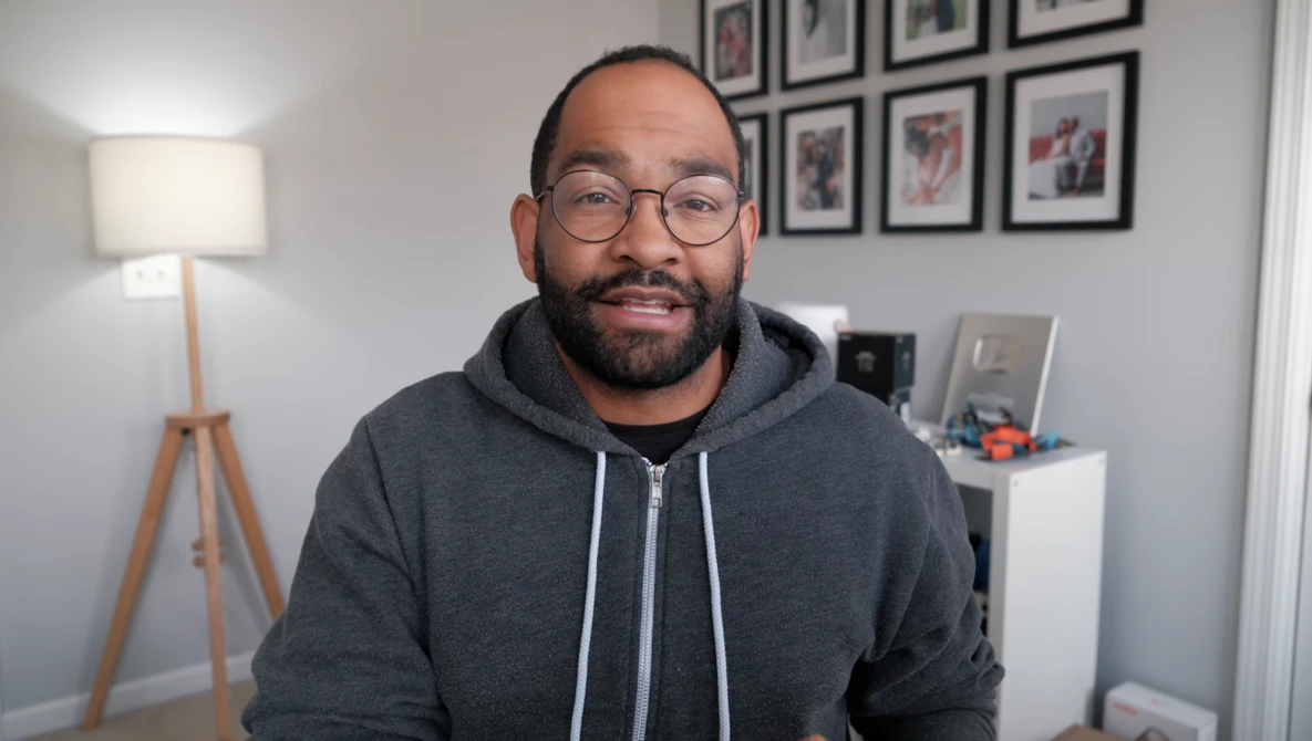 Man wearing glasses and dark hoodie smiling at camera in home office setting.