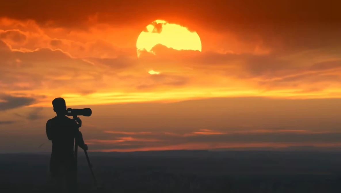 Silhouetted photographer with telephoto lens on tripod shooting a massive golden sunset.