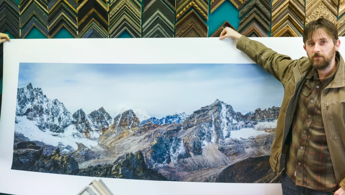 Photographer holding a large alpine landscape print featuring snow-capped peaks and rocky terrain.