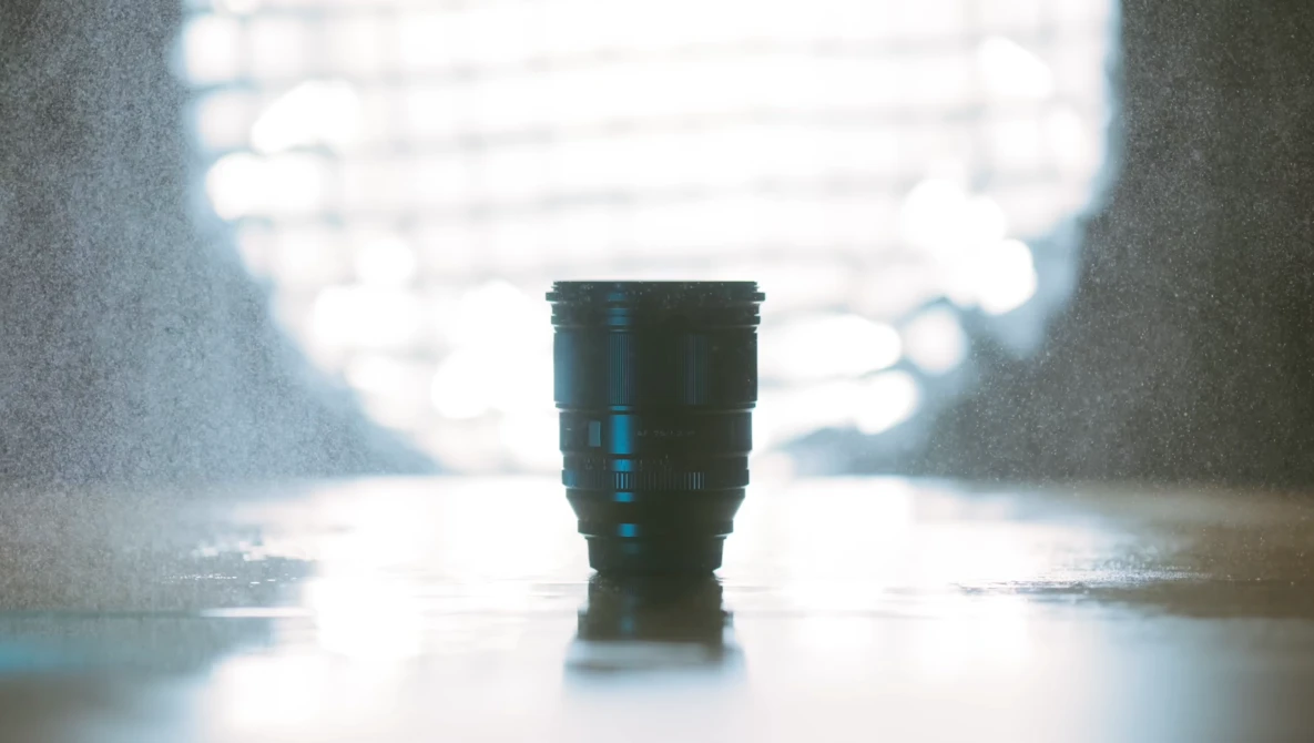 Dark telephoto lens standing upright on wet pavement with blurred bright background.