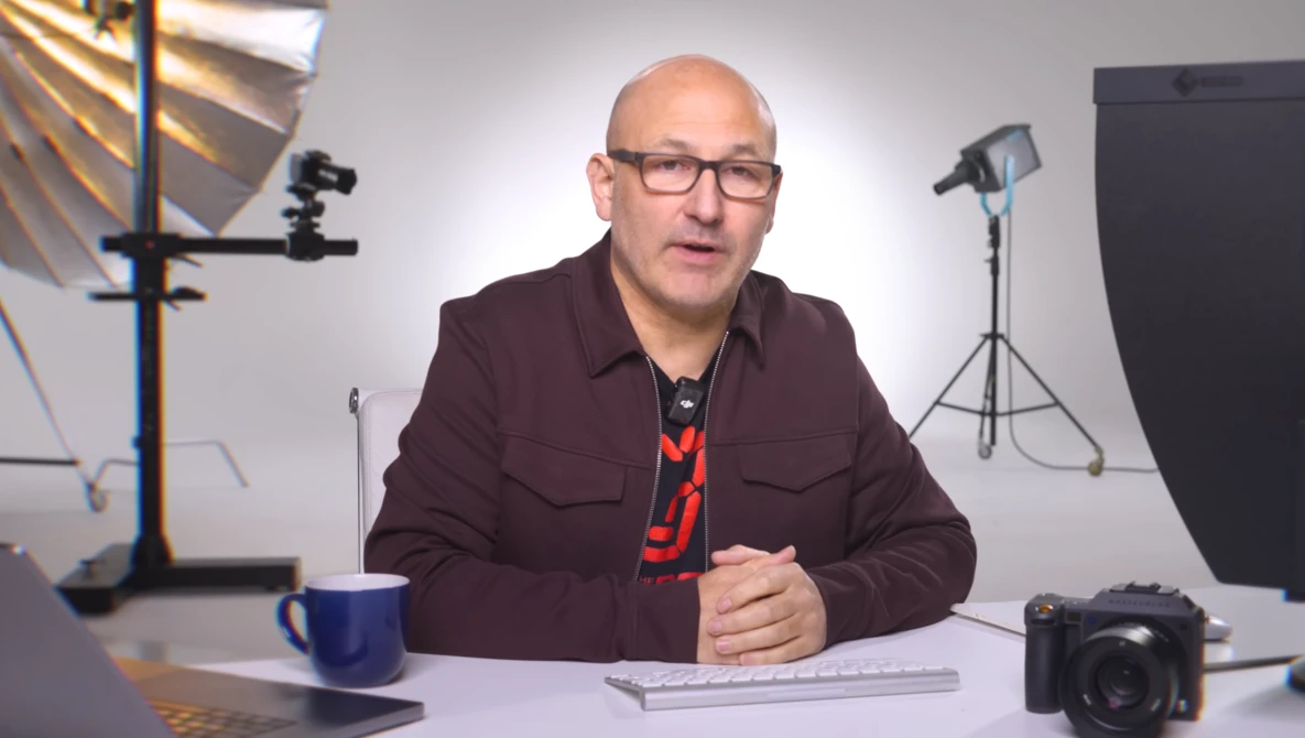 Man in maroon jacket speaking at desk in professional studio with lighting equipment.