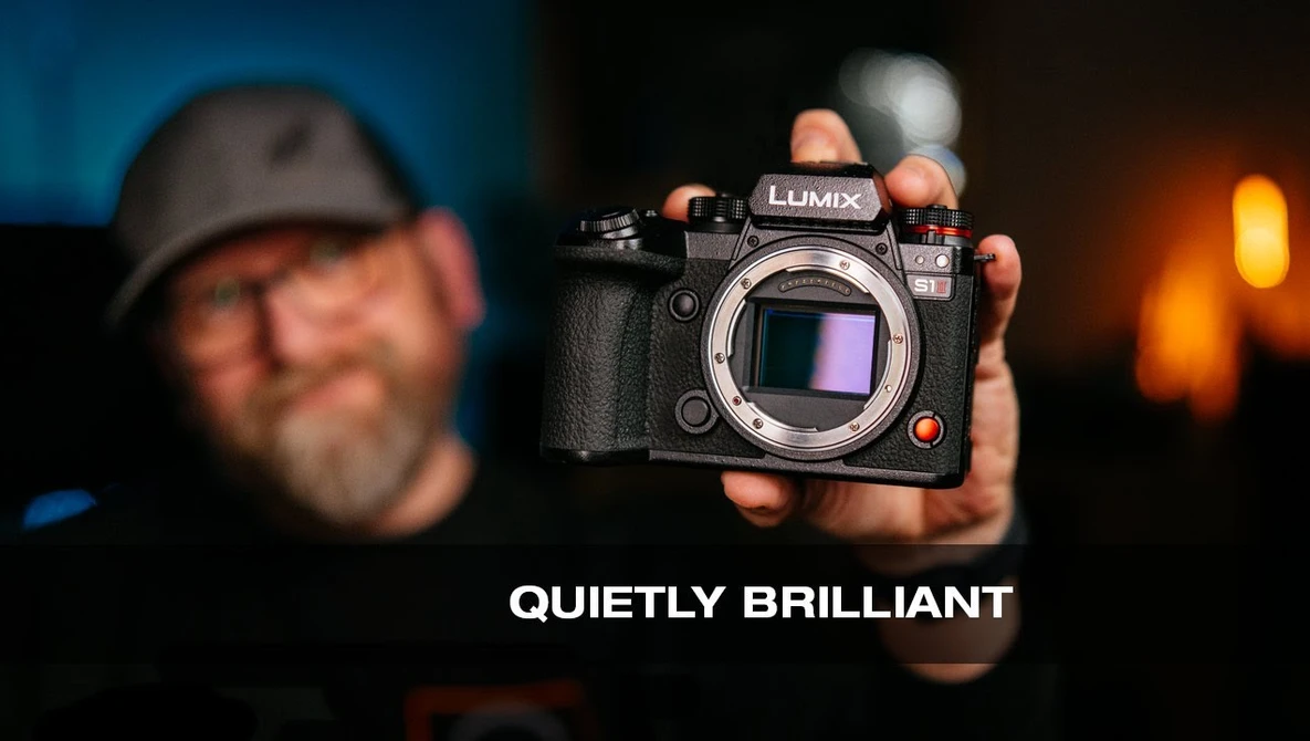 Photographer holding a Lumix mirrorless camera body toward the camera against a blurred background with warm lighting.