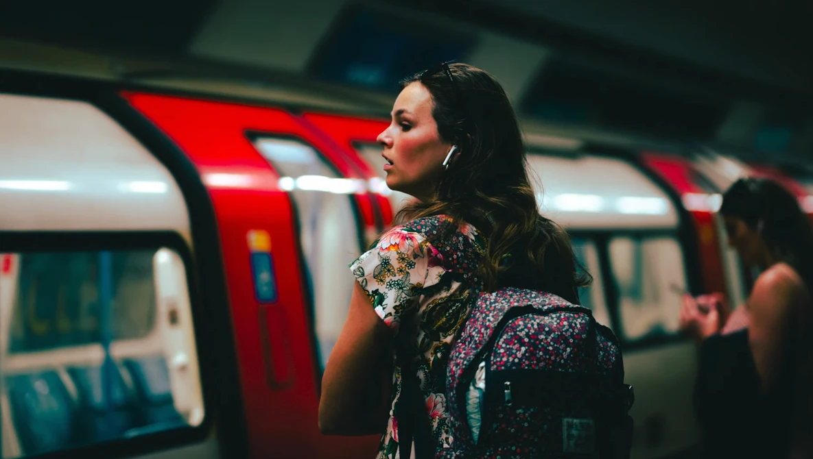 Woman with dark hair wearing a colorful floral jacket stands in a subway car with red doors and blue seating visible behind her.