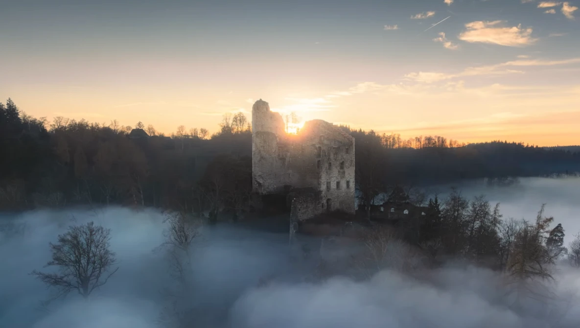 Medieval stone tower backlit by sunrise, surrounded by dense fog over a forested landscape.