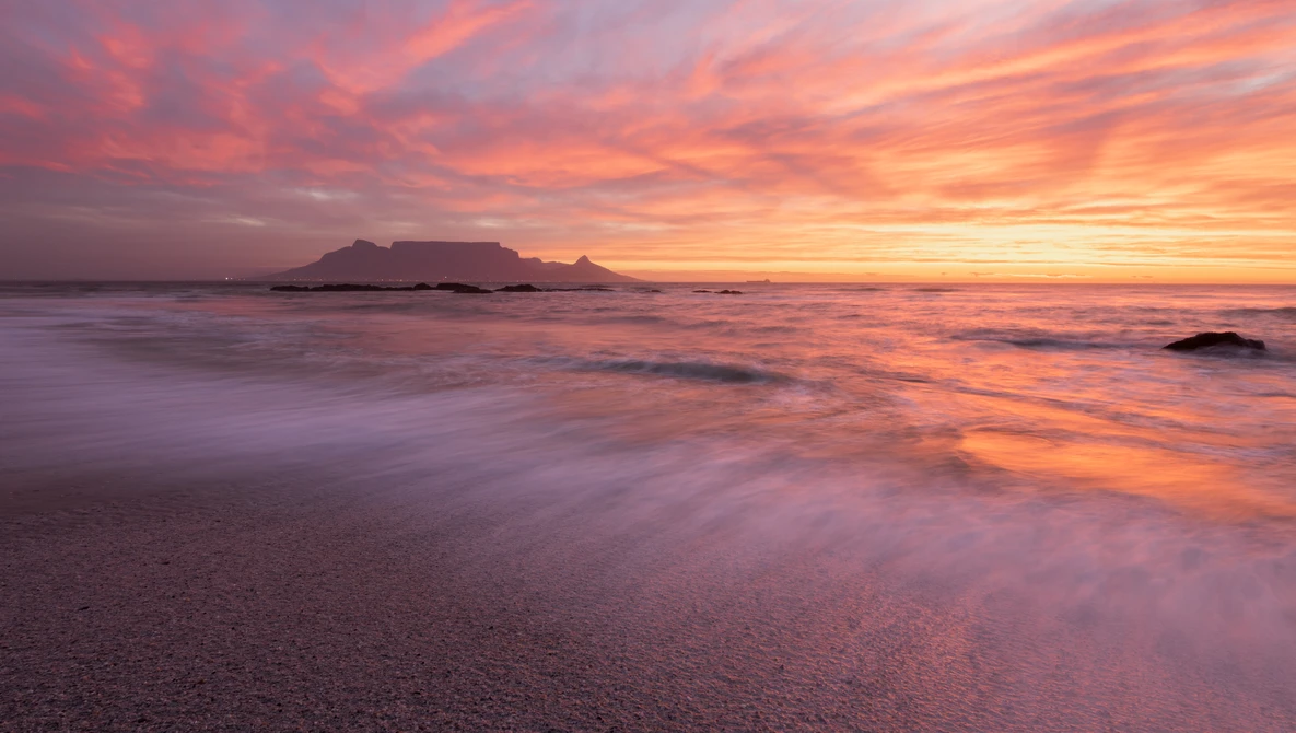 Sunrise over a coastal bay with a flat-topped mountain silhouetted against an orange and pink sky, waves washing onto the beach in the foreground.