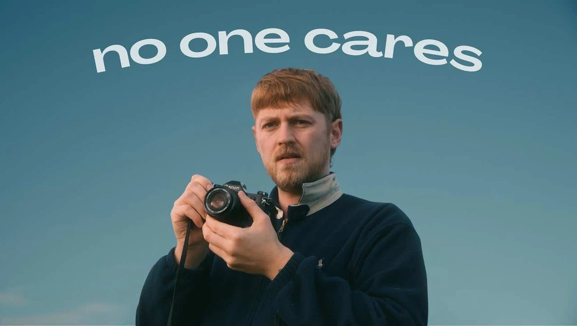 Man holding a vintage camera against a blue sky backdrop with "no one cares" text overhead.