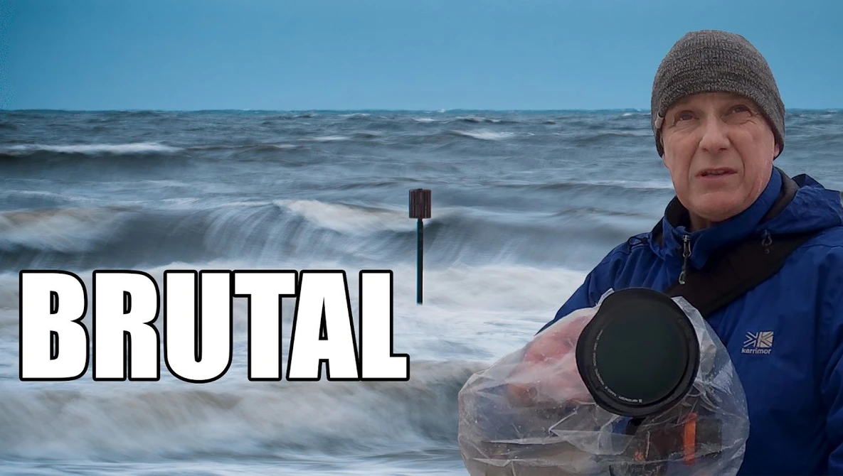 Photographer in winter gear holding waterproof camera equipment by the ocean.