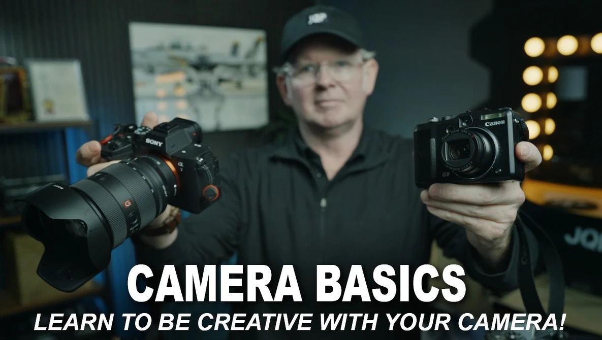 Photography instructor holding a mirrorless camera and compact camera while demonstrating equipment in a studio setting.