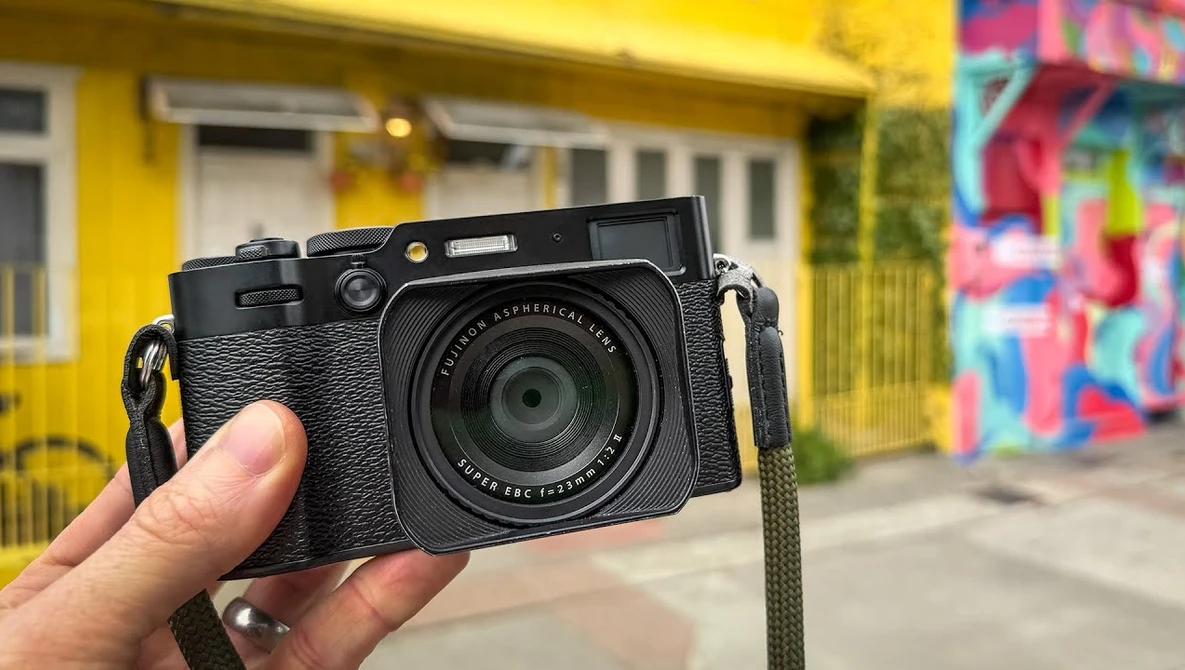 Black rangefinder camera with textured grip held in hand against urban backdrop.