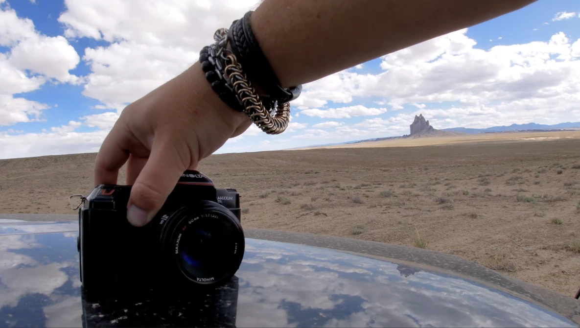 Hand holding a compact camera over a shallow water pool in an arid landscape with rock formations.
