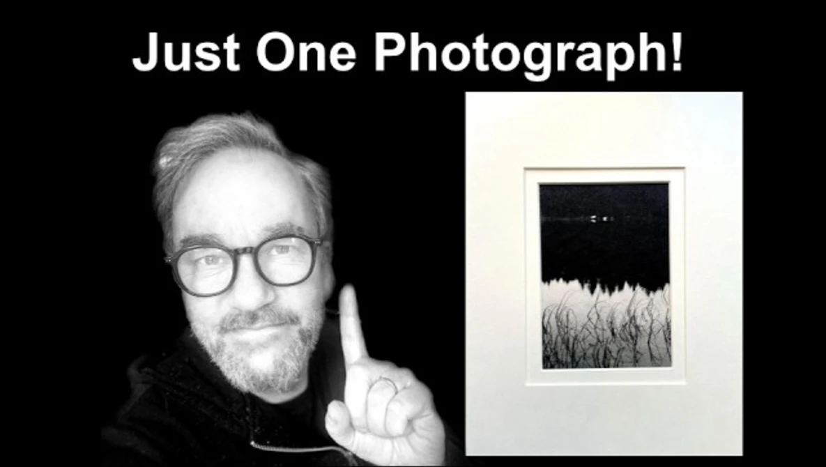 Photographer holding up one framed black and white landscape photograph featuring a dark sky above a textured field.