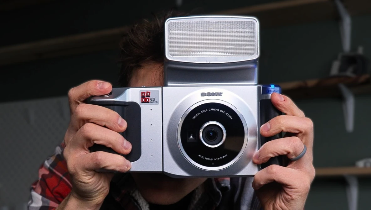 Hands holding a white and black instant film camera with a flip-up flash attachment.