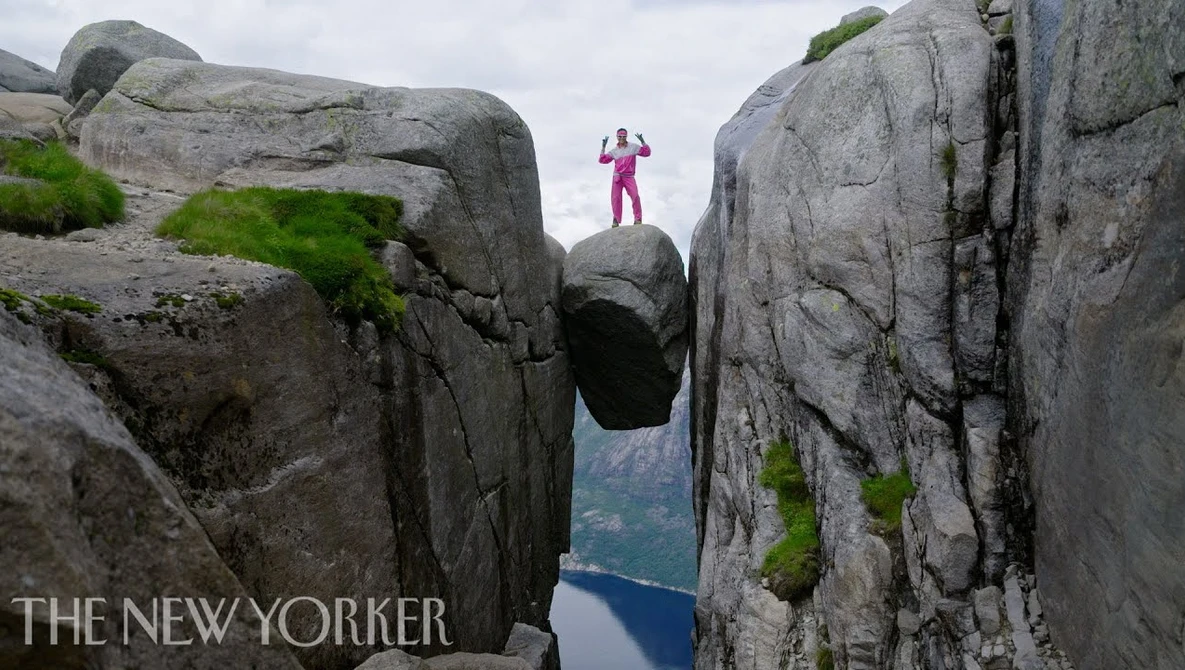 Person in magenta jacket standing on narrow rock outcrop between two steep granite cliffs overlooking a fjord.